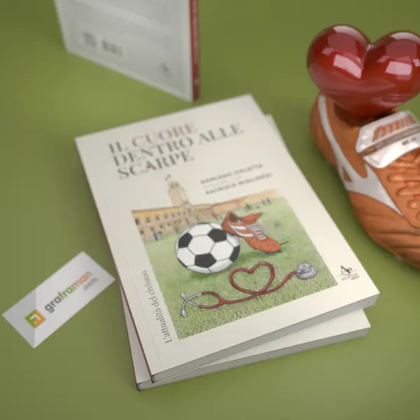 Vista dall'alto di due copie sovrapposte del libro «Il cuore dentro alle scarpe», con l'illustrazione di copertina in evidenza e un cuore rosso lucido appoggiato su una scarpa da calcio - Graframan