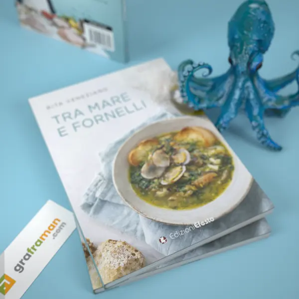 Vista dall'alto di due copie sovrapposte del libro «Tra mare e fornelli», con la copertina fotografica del piatto di vongole e crostini e il polpo blu in appoggio sul libro - Graframan