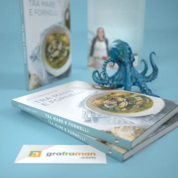 Dettaglio dei dorsi azzurri di due copie del libro «Tra mare e fornelli», con titolo e nome dell'autrice, e la copertina fotografica della copia in alto - Graframan
